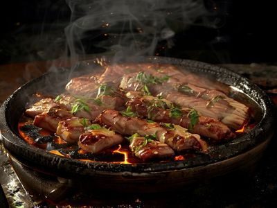 韩国烤五花肉在炭火烤架上滋滋作响，油脂滴落产生烟雾，旁边配有生菜包裹材料，氛围感十足