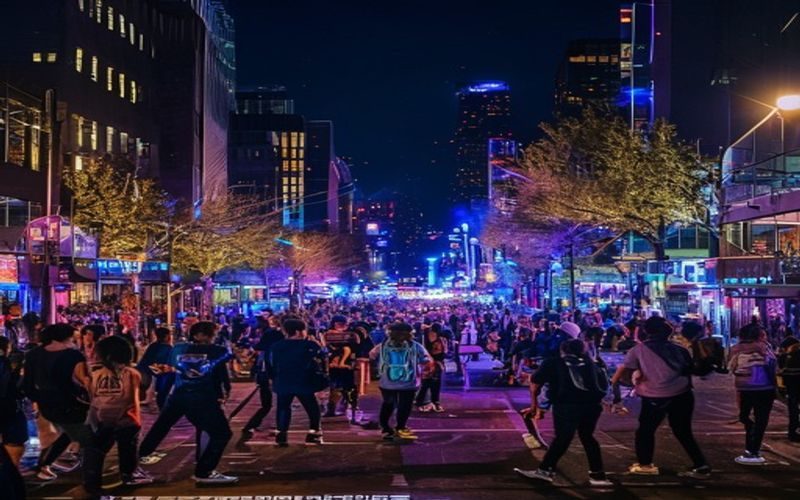 首尔弘大街区夜晚霓虹灯闪烁，街头艺人表演吸引大批年轻人围观，画面充满活力与潮流气息